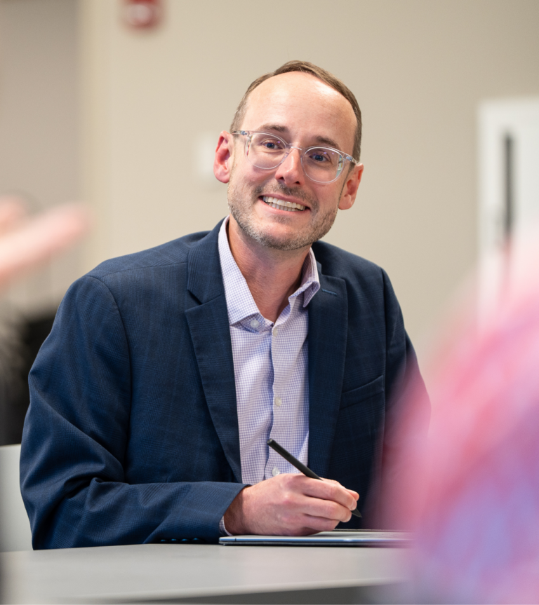 Business professional wearing a navy blazer and clear glasses, smiling while seated at a table during a meeting.