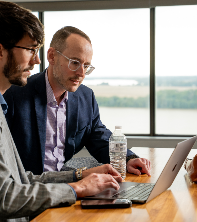 Two colleagues in business attire review work together on a laptop at a conference table, collaborating in a bright office with large windows.