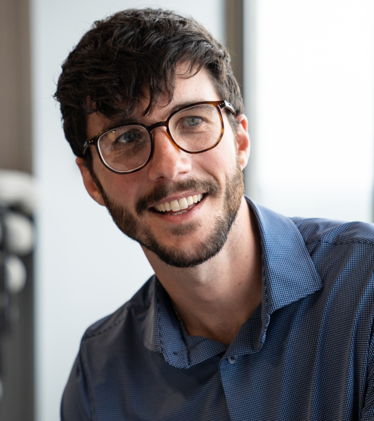 Close-up portrait of a smiling business professional wearing glasses and a blue patterned shirt in a bright indoor setting.